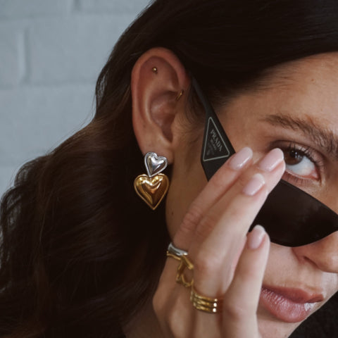 Woman wearing two tone gold and silver heart earrings and rings, holding prada sunglasses