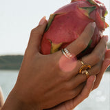 Person holding a dragon fruit by a body of water wearing freshwater pearl rings and waterproof gold rings