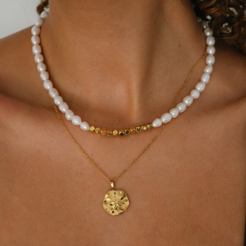 Close-up of a person wearing a gold sea dollar pendant necklace with a pearl necklace layer