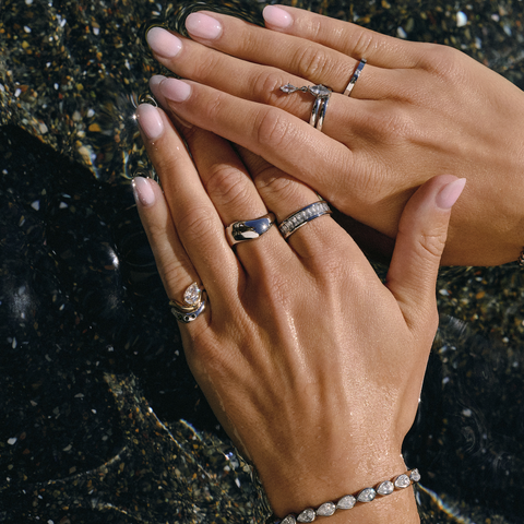 Two hands with manicured nails resting on a dark, wet stone surface, showcasing a collection of silver-toned jewelry including tennis bracelets and rings with clear gemstones.