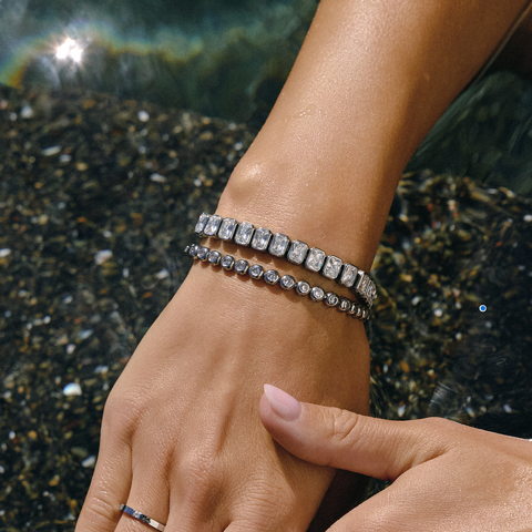 A close-up of a person's wrist wearing two silver-toned bracelets, one with rectangular clear stones and the other with round clear stones, set against a dark pebble-and-water background.