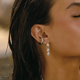 A close-up lifestyle shot of a woman with wet hair wearing a silver hoop earring stacked with clear faceted gemstones and a matching drop earring, set against a softly blurred outdoor background.