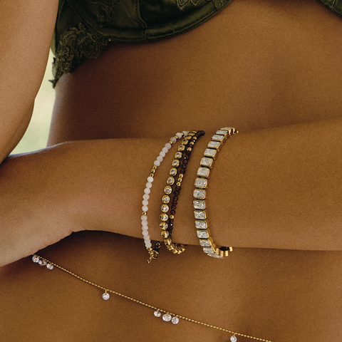 A close-up lifestyle shot of a model's wrist adorned with multiple stacked bracelets, including a gold tennis bracelet with clear stones, a gold bracelet with dark brown beads, a gold bracelet with white beads, and a delicate gold chain anklet with clear rhinestones resting on her hip.