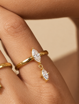 A close-up of a gold ring with two dangling marquise-cut clear stones on a person's finger, shown against a warm beige background.
