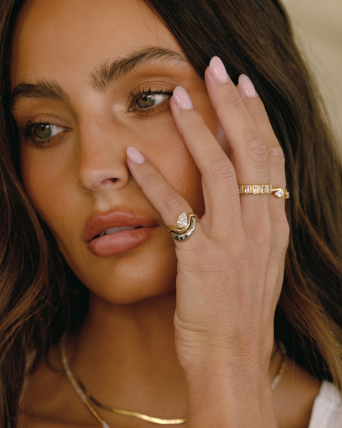 A close-up lifestyle shot of a woman with long brown hair wearing multiple gold rings, including one with a large clear gemstone and another with small clear stones, along with layered gold and silver necklaces.