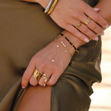 A close-up lifestyle shot of a model's hands wearing an assortment of gold jewelry, including rings, a delicate chain hand bracelet, and a beaded bracelet, styled with an olive green dress.